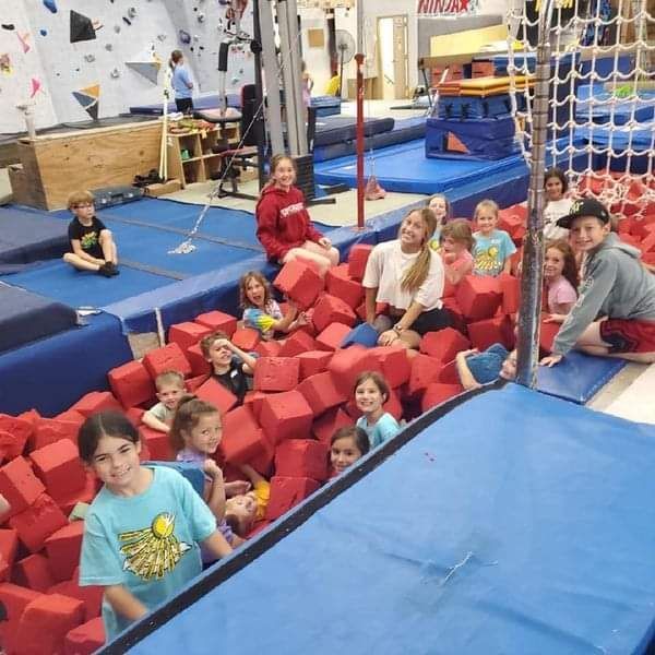 Group of kids smiling and playing in a pit of red foam cubes at a gym.
