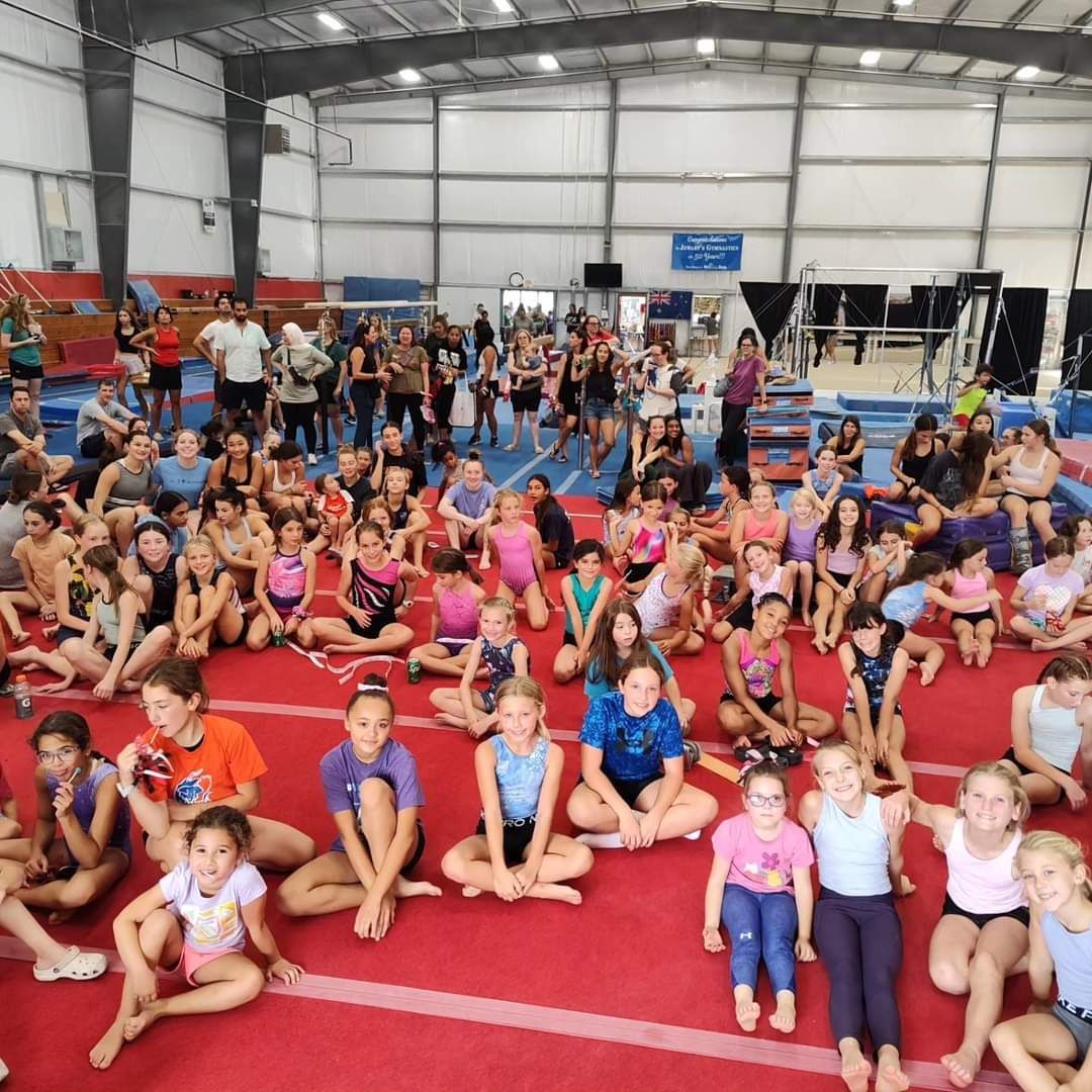 Large group of children and adults in a gymnastics gym, sitting on a red mat.