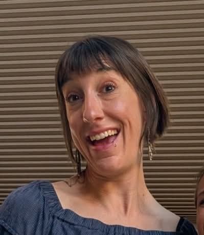 Woman with short brown hair and bangs, smiling, wearing earrings and a blue top, against a striped background.