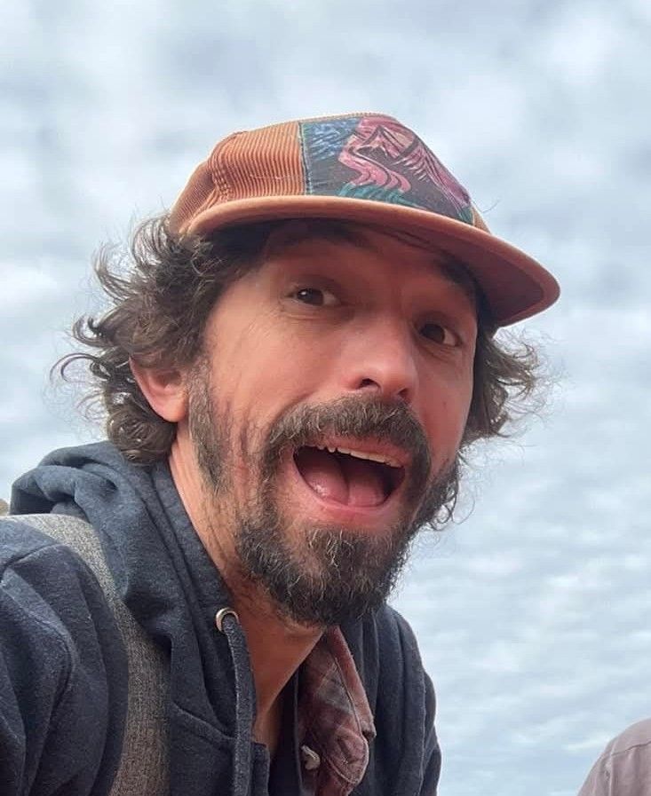 Man with curly hair, open mouth, wearing a hat, looking at camera.