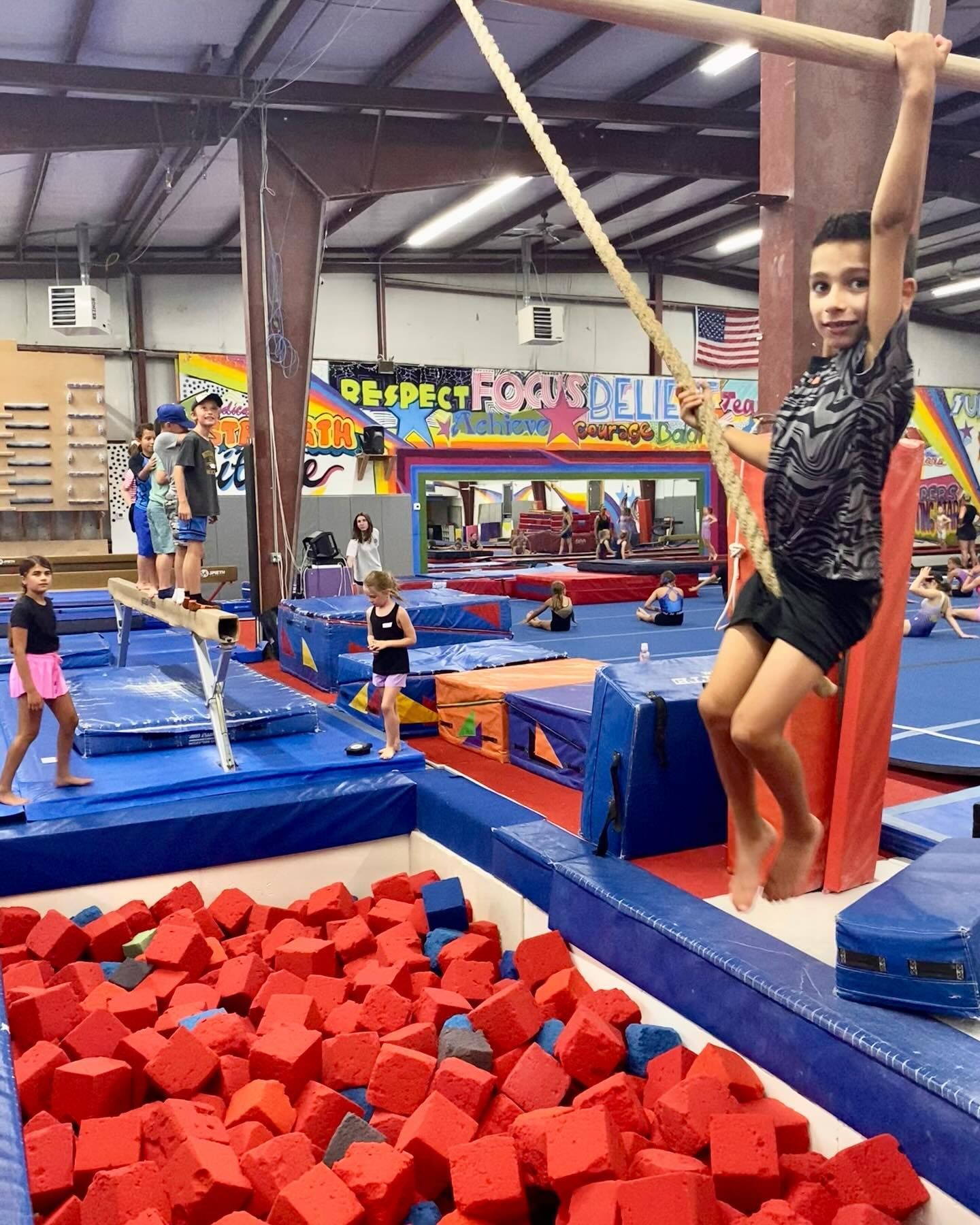 Boy swinging on a rope in a gymnastics gym with foam pit. Others on equipment in background.