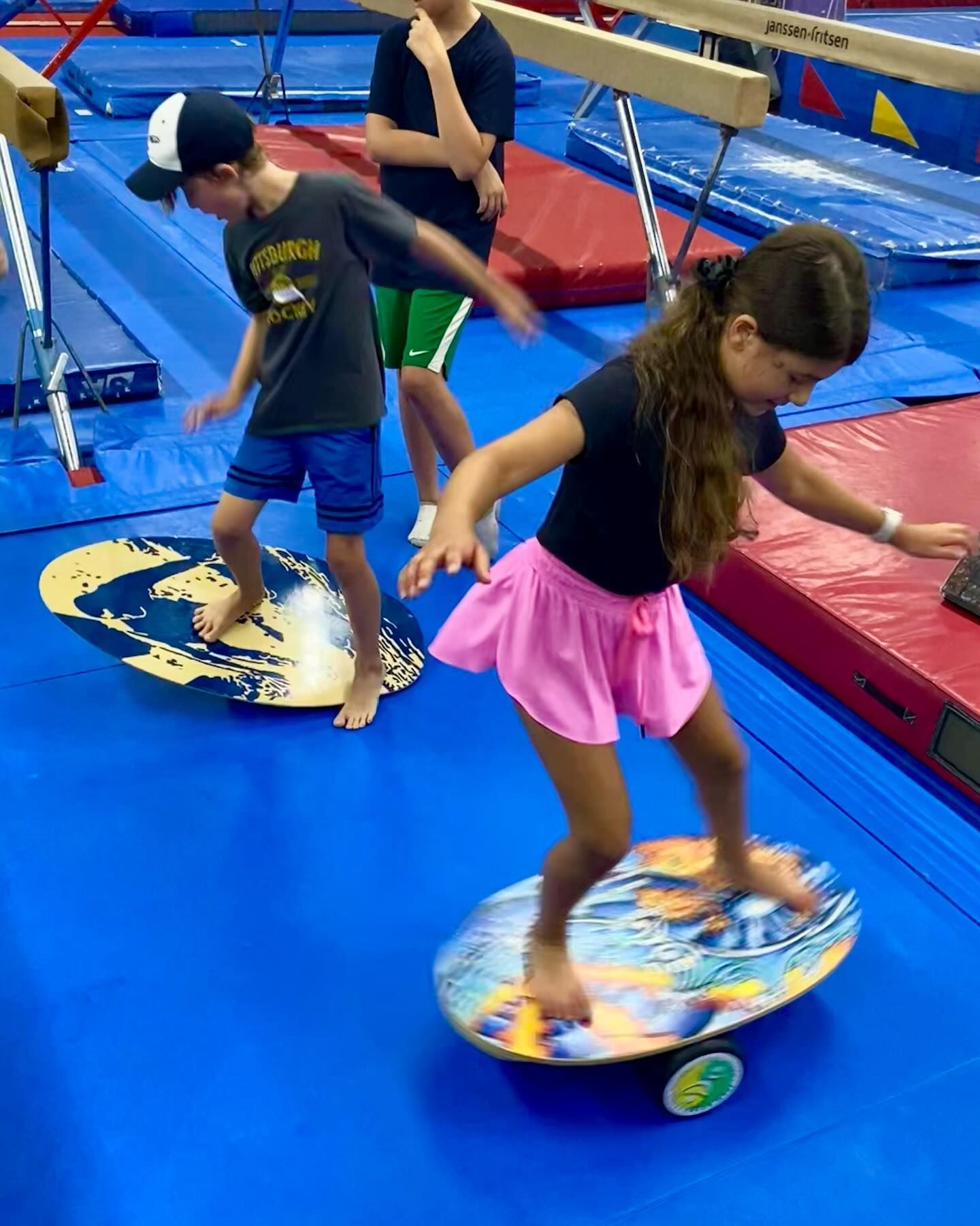 Children balancing on wobble boards in a gym with blue mats and balance beams.
