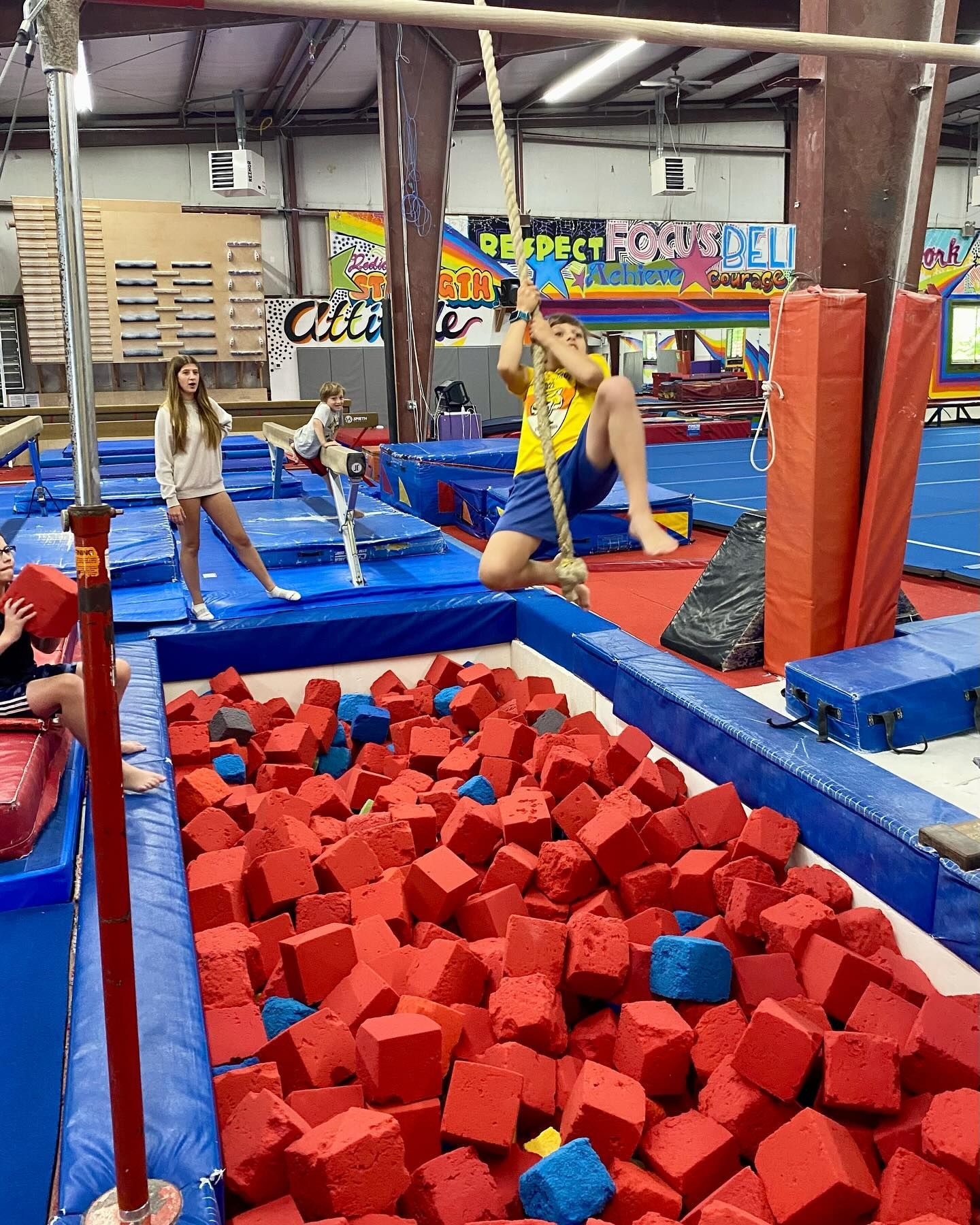 Gymnast swinging on a rope, landing in a foam pit. Others watch in a gym setting.