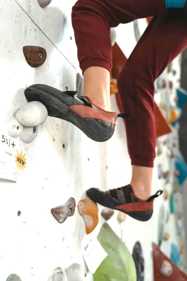 Rock climber's feet in specialized shoes on a climbing wall, red pants, hands out of frame.