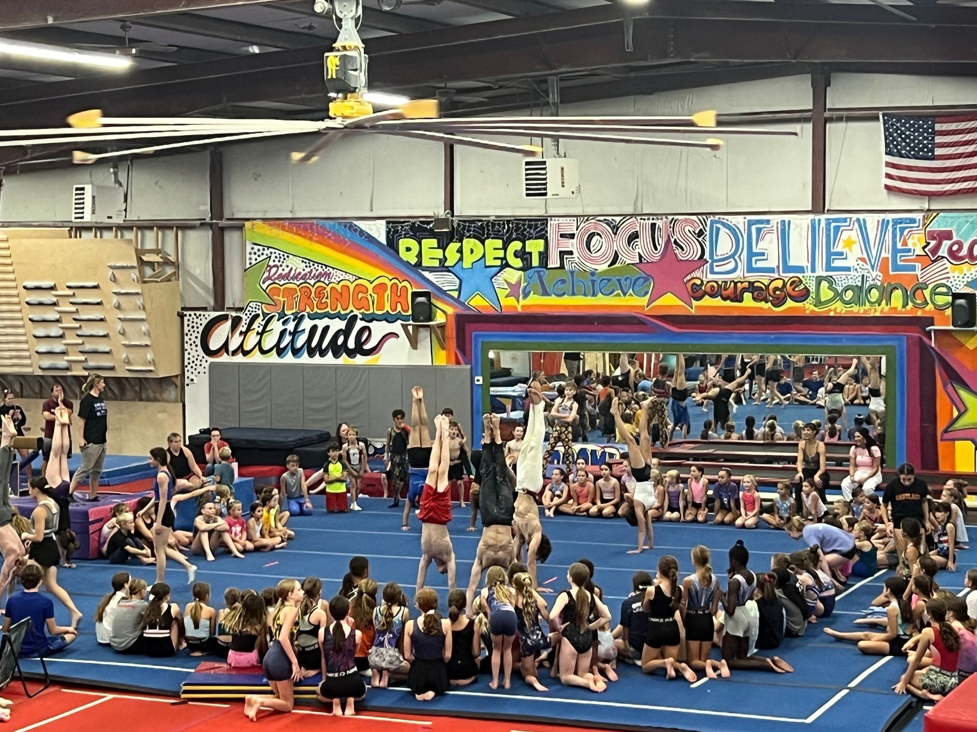 Gymnastics class in a colorful gym; children watch others perform handstands and poses.