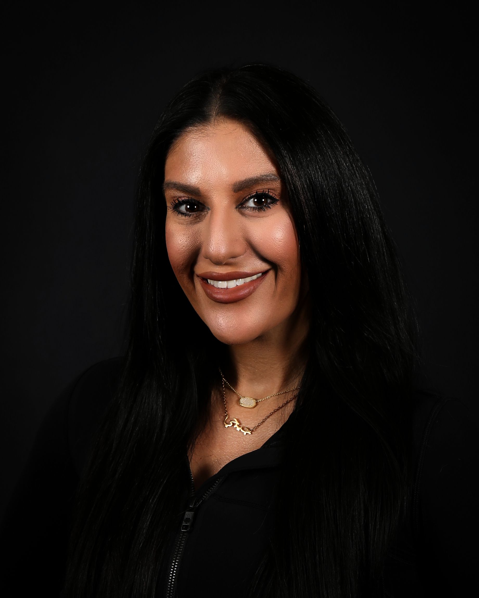 Woman with long dark hair smiles, wearing a black top and gold necklaces, set against a dark background.