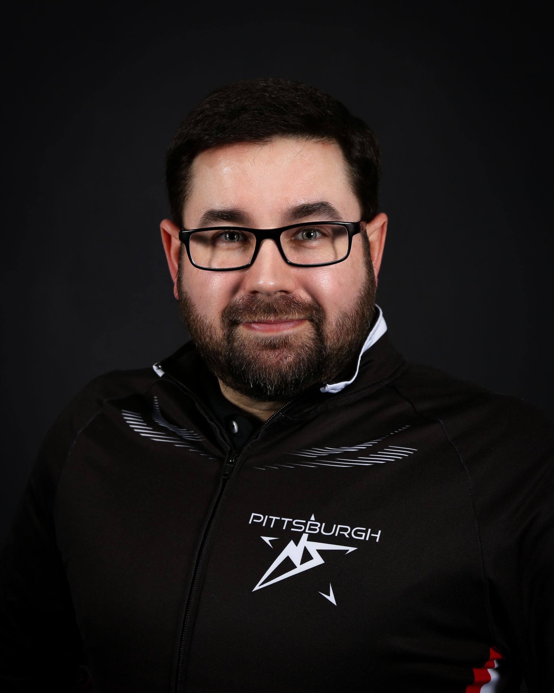 Man with beard and glasses, wearing a black and white Pittsburgh jacket, smiling against a dark background.