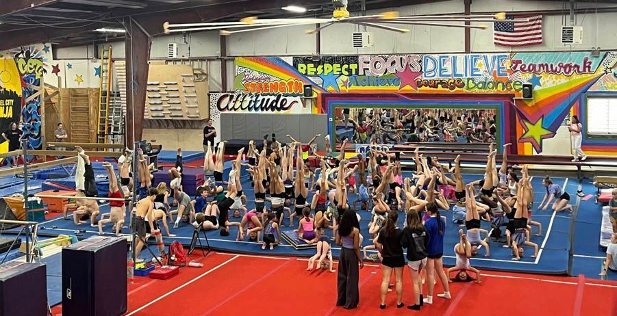 Gymnastics class in large gym, with many people doing handstands and stretching. Bright mural background.