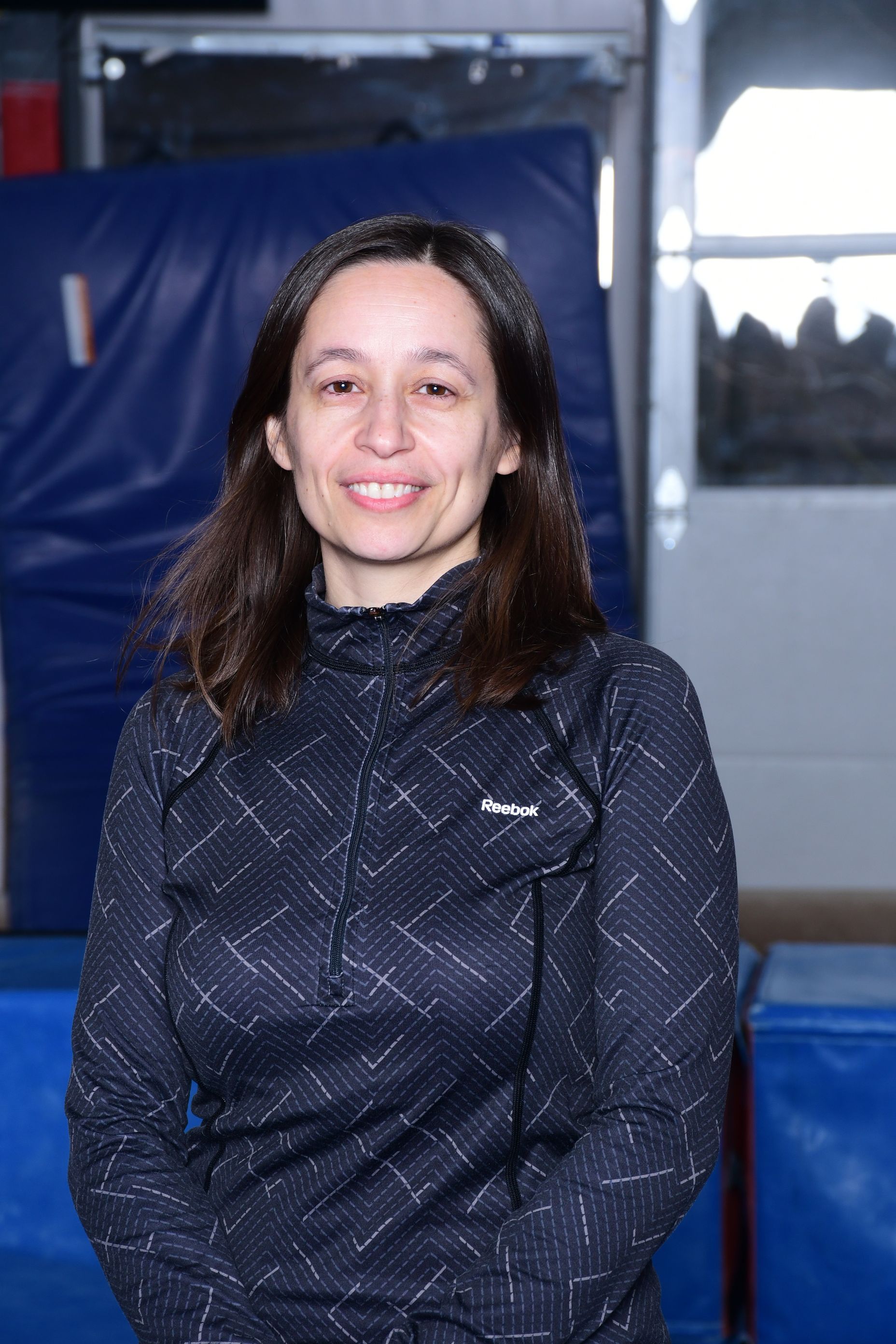 Woman smiling, wearing a dark patterned athletic top, standing in a gym with blue mats.