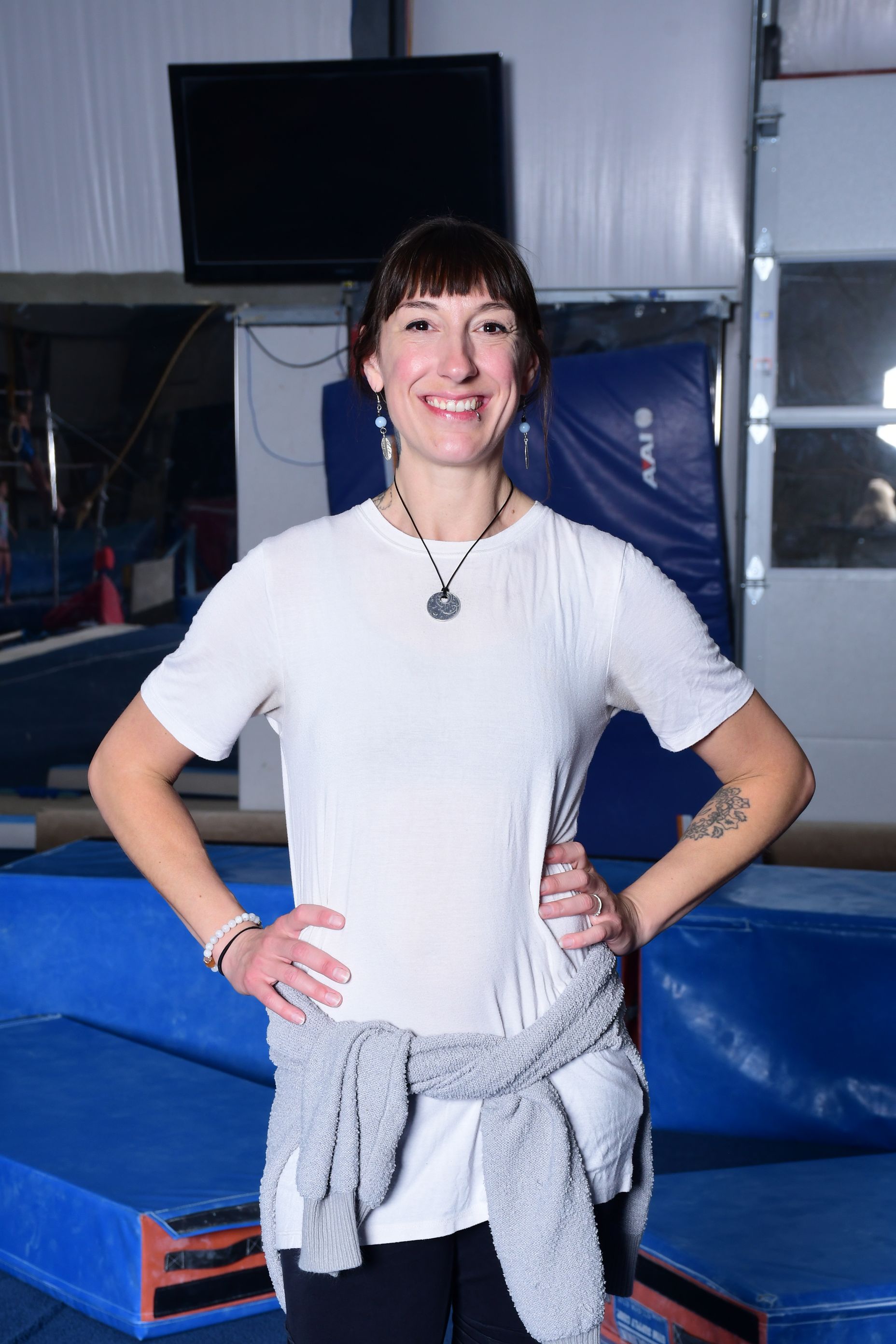 Woman in a white shirt and black pants, smiling with hands on hips, in a gym with blue mats.
