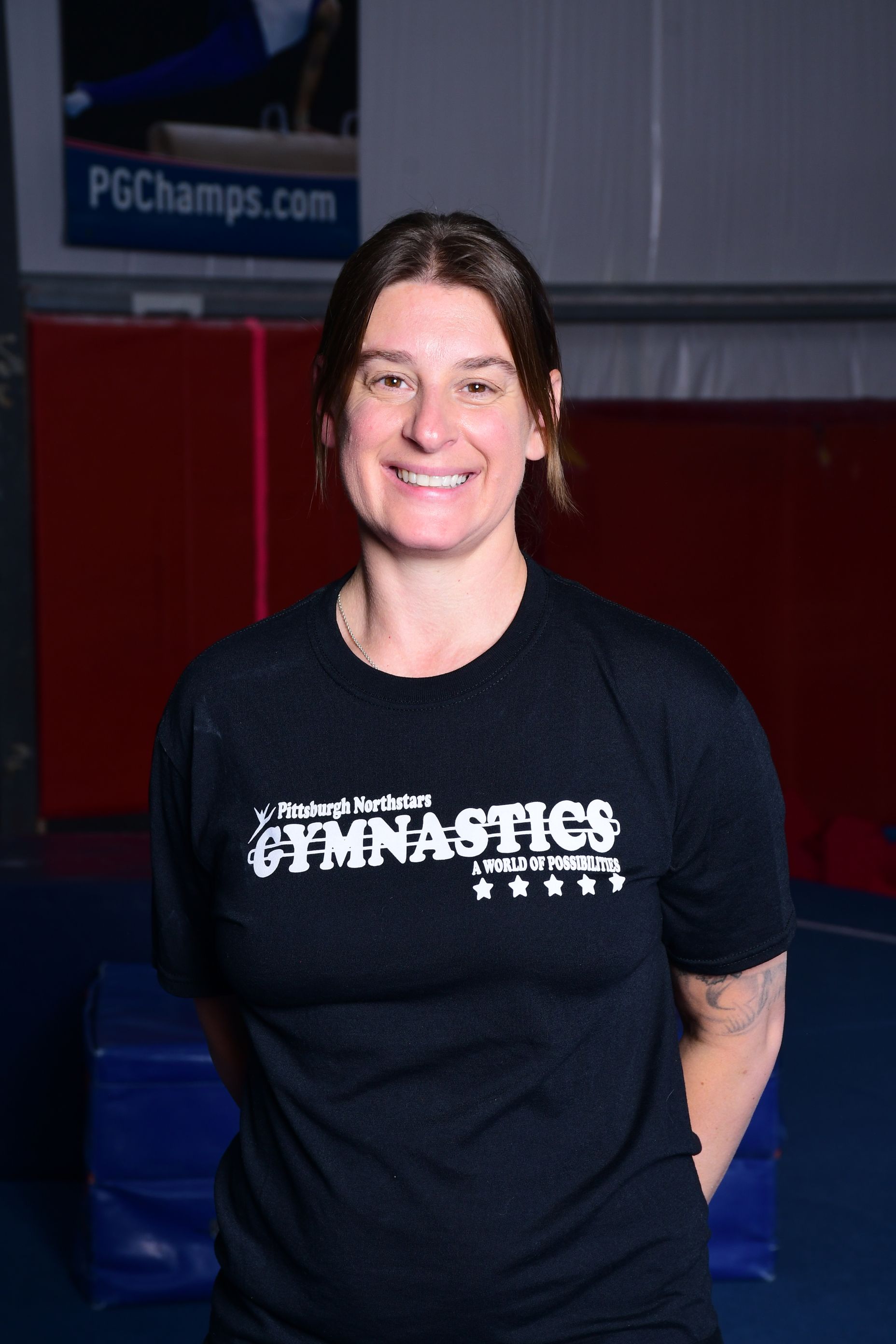 Woman with short brown hair, smiling, wearing a black gymnastics t-shirt, standing in a gym.