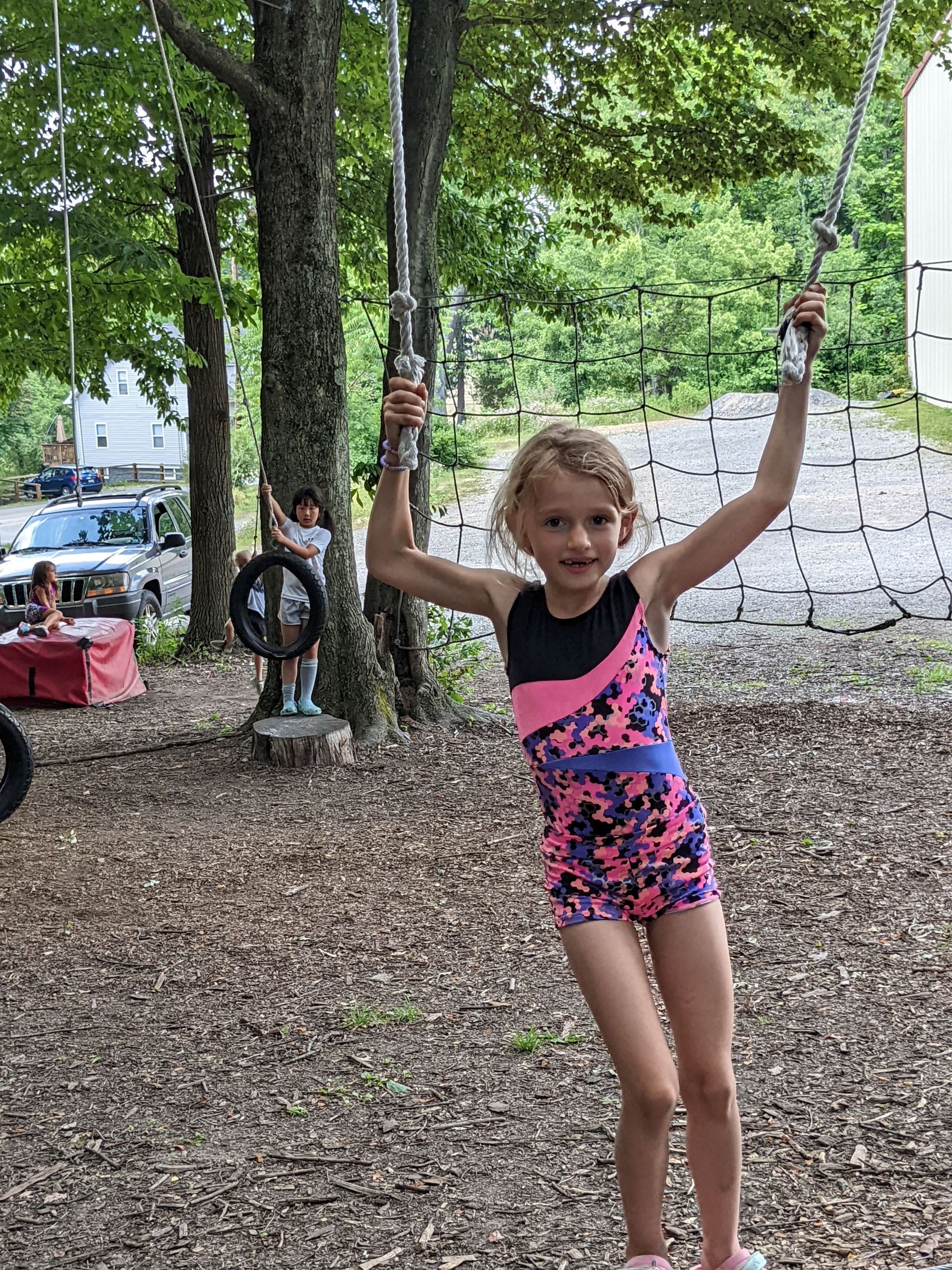 Girl in gymnastics leotard on rope swing outdoors, smiling with arms raised.