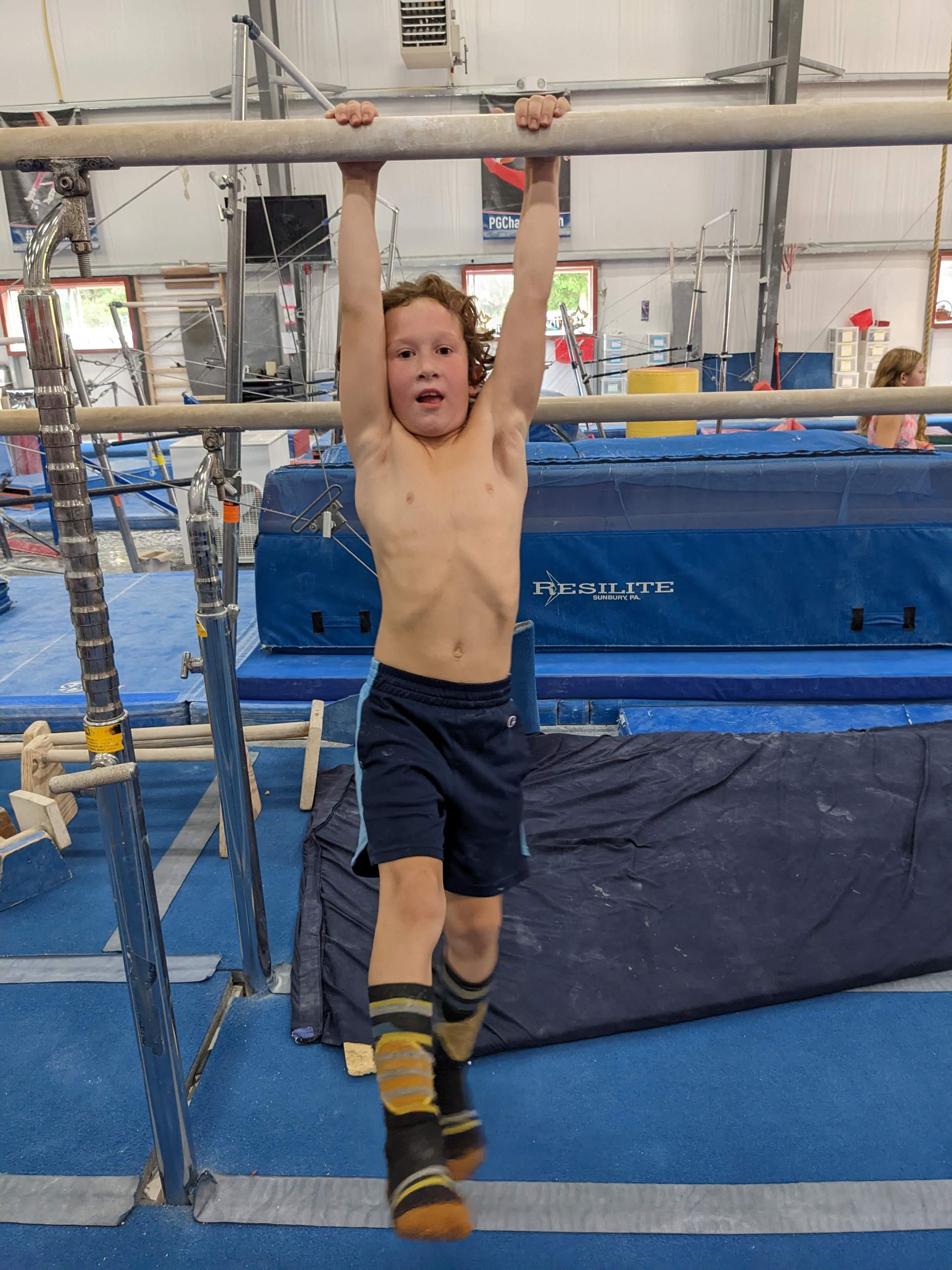 Boy hangs from a horizontal bar in a gym, wearing shorts and socks, looking determined.