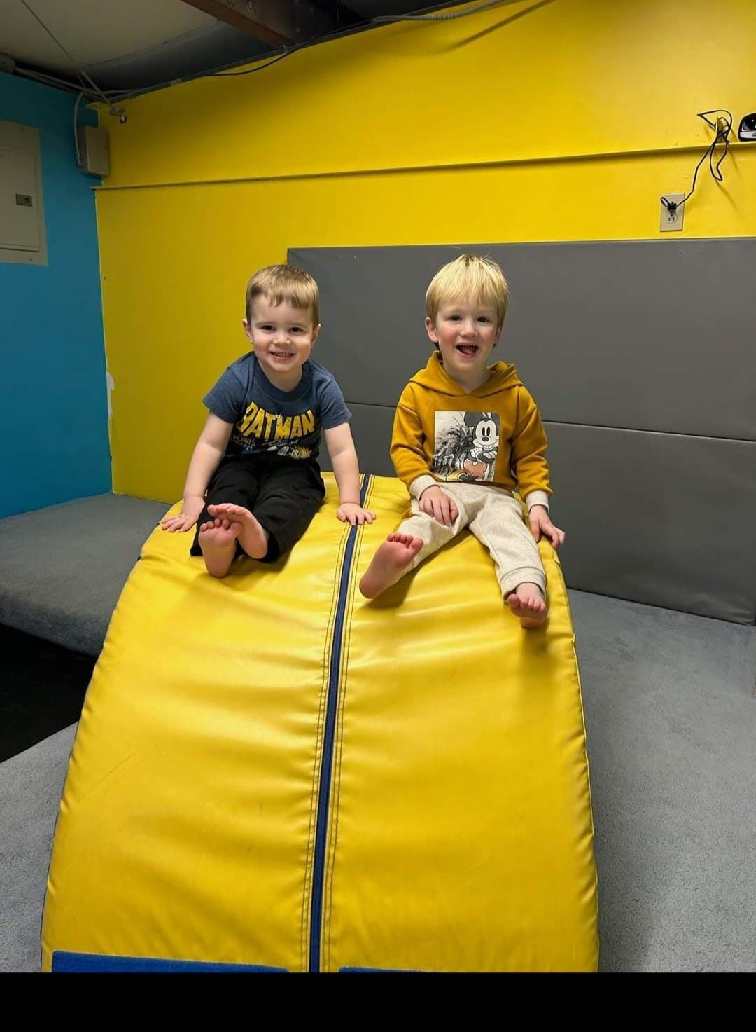 Two young boys smiling, sitting on a yellow padded obstacle in a gym; yellow and blue walls.