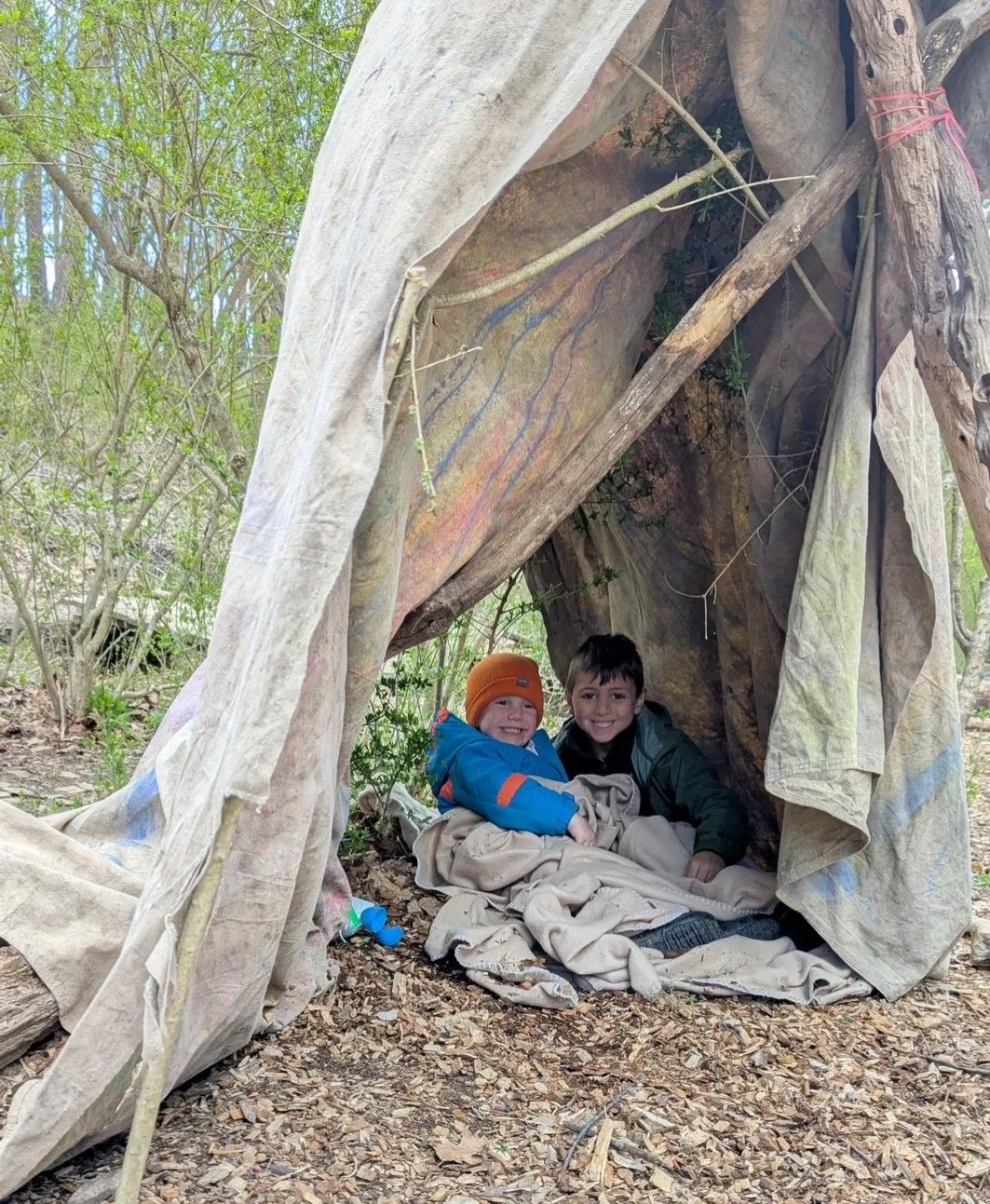 Two children smile inside a rustic fort made of branches and fabric in a wooded area.