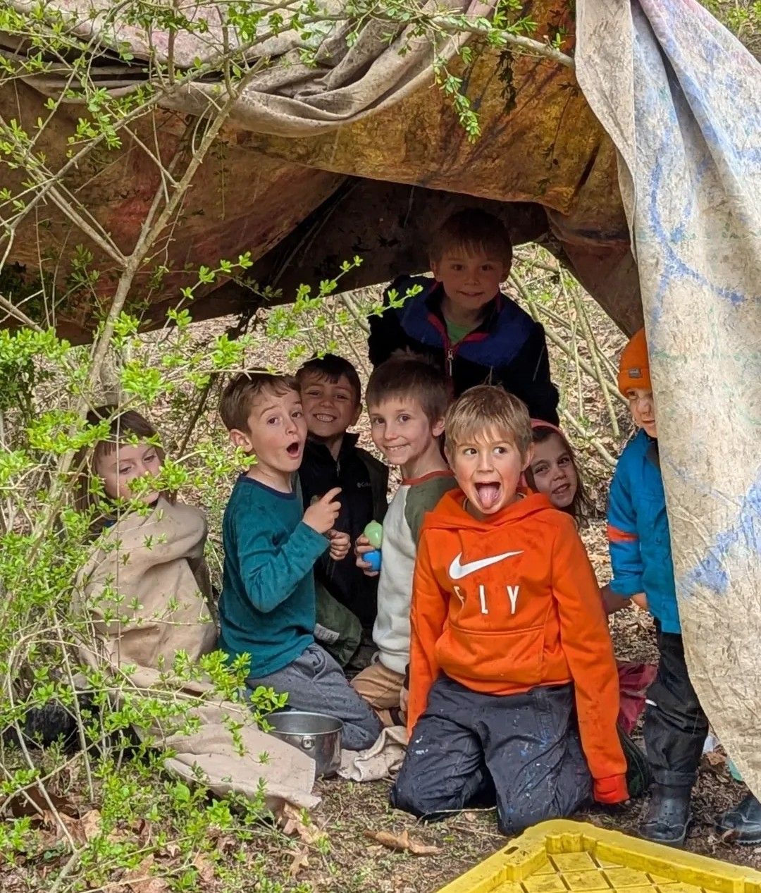 Group of children in a makeshift tent outdoors, smiling with joyful expressions.