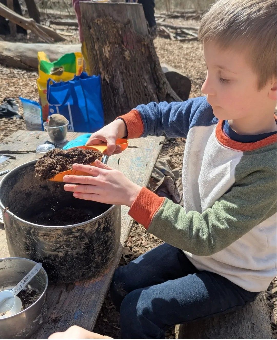 A young boy using a small orange shovel to move dirt into a large metal pot in an outdoor setting.