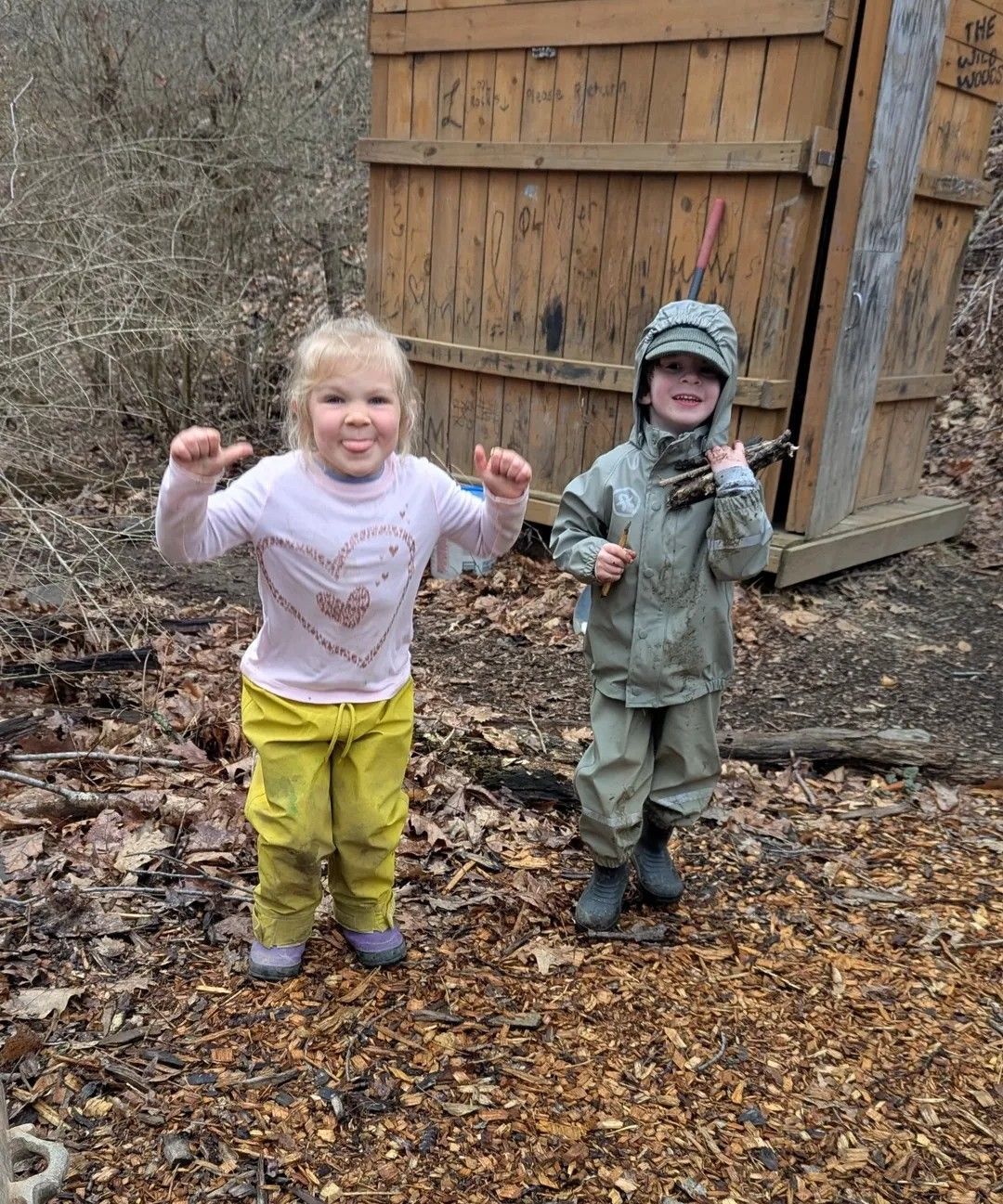 Two young children playing outside by a wooden structure; one in rain gear, both smiling.