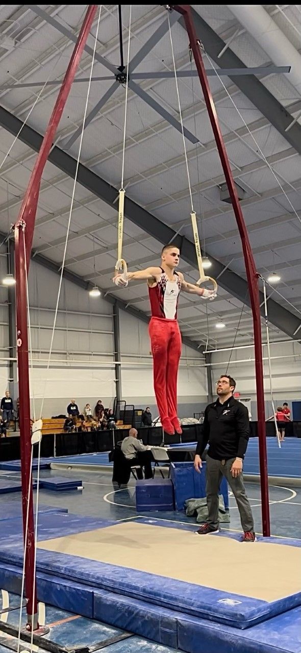 Gymnast on rings in red and white uniform, performing a routine. Coach watches nearby.