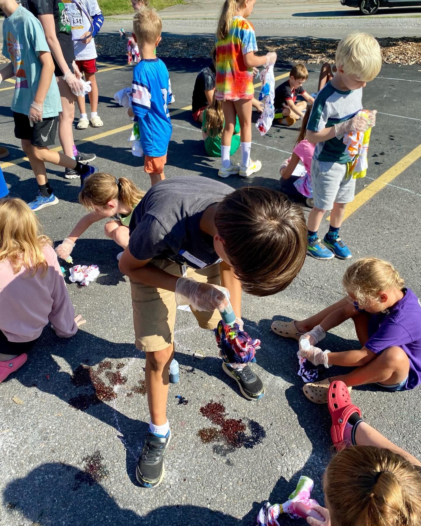 Children tie-dyeing clothes on asphalt. They wear gloves, and their clothes are stained with dye.