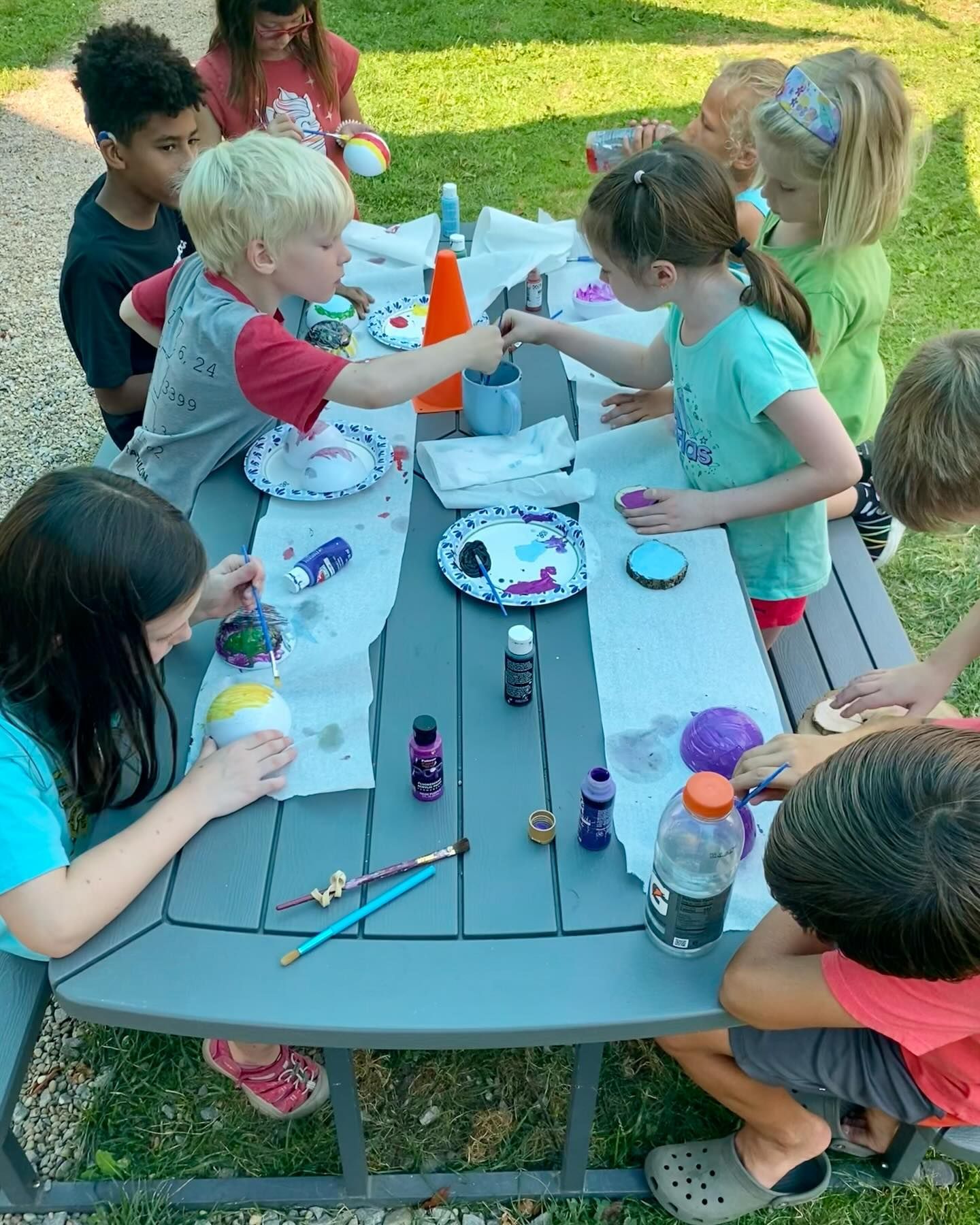 Children painting ornaments at an outdoor picnic table. They are using paintbrushes and various colors.