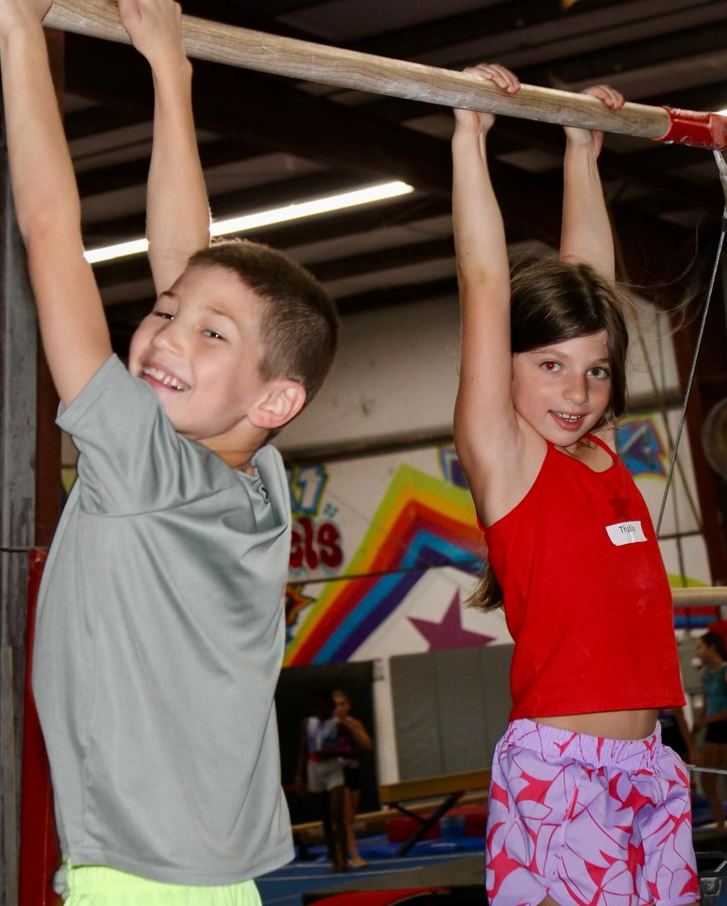 Two children hanging from a bar, smiling. Boy in gray, girl in red, in a gym.
