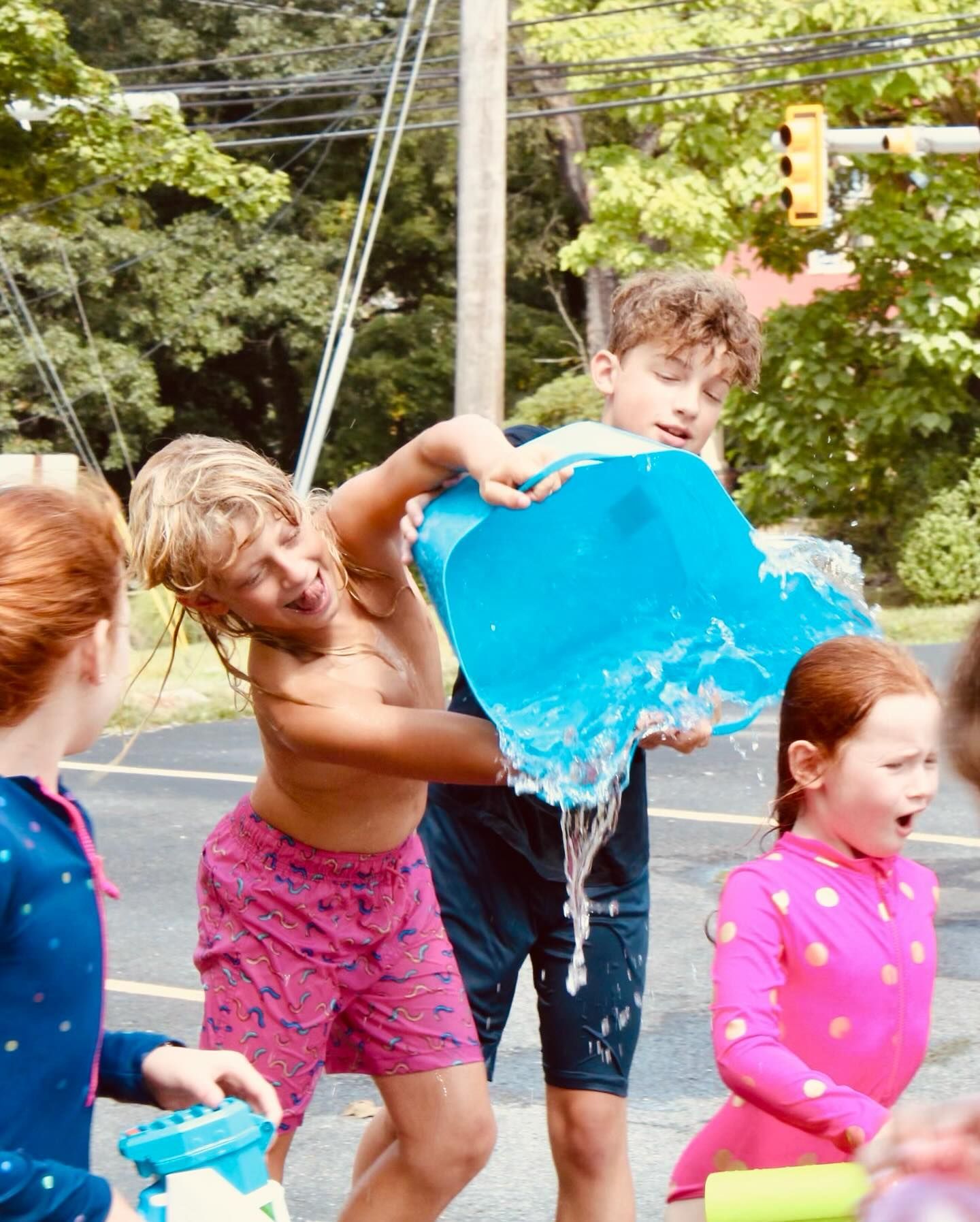 Children playing with water. Boy douses another with blue bucket. Others watch. Bright summer day.