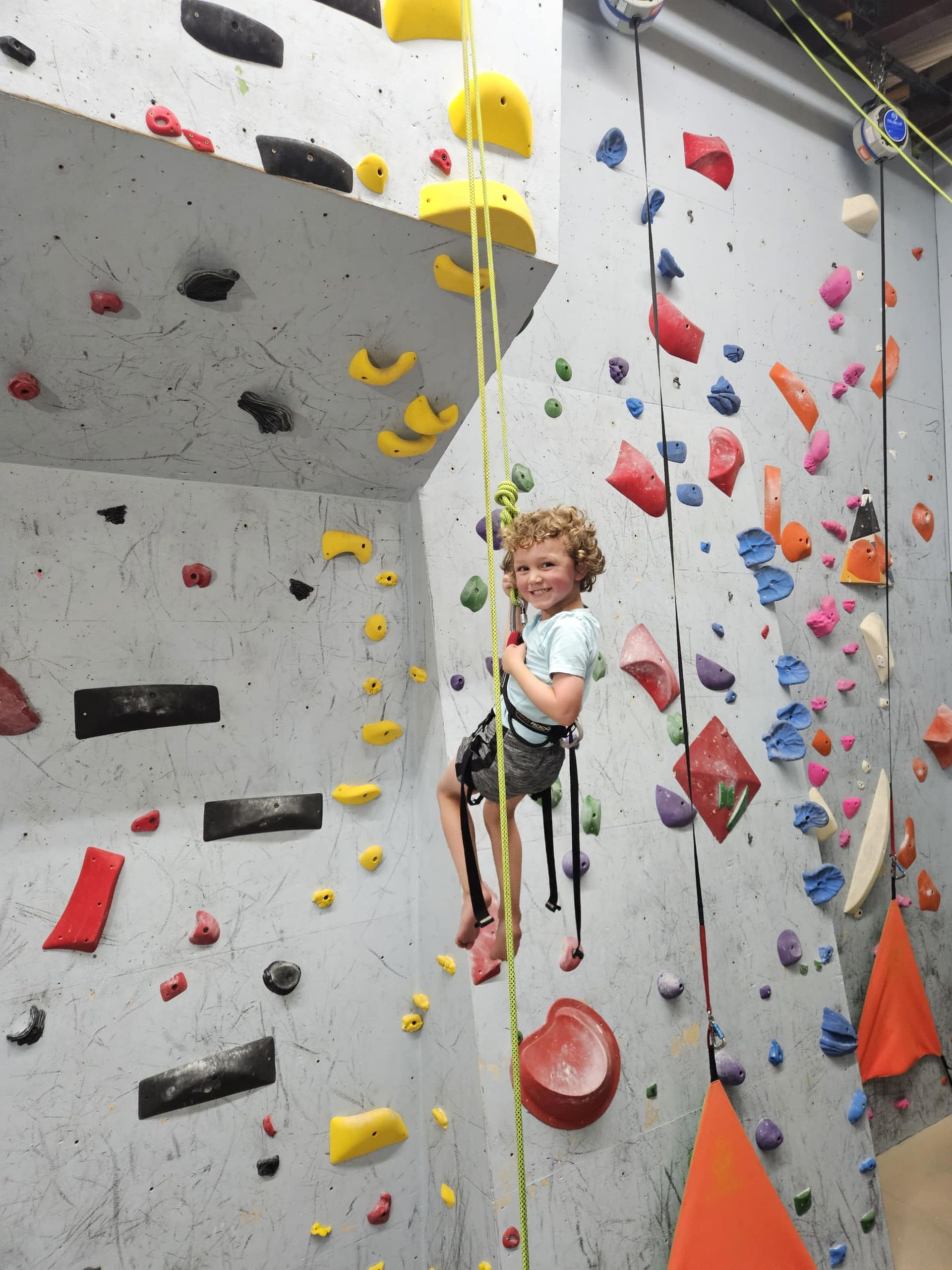 Young child smiling while rock climbing on a colorful indoor wall, secured with rope and harness.