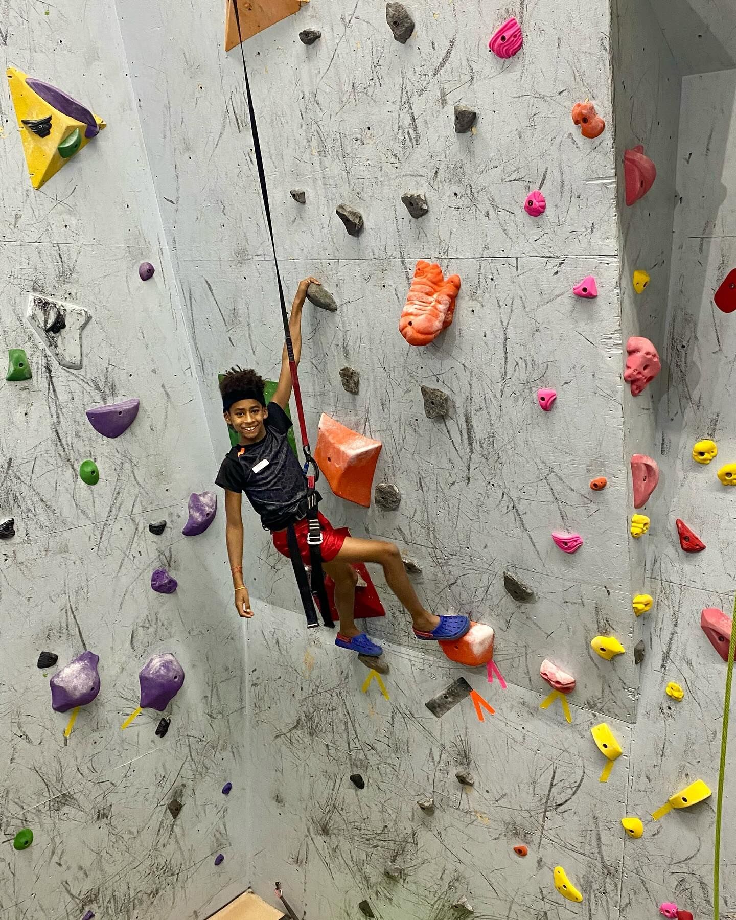 Young person rock climbing, secured with a harness, smiling, on a textured wall with colorful holds.