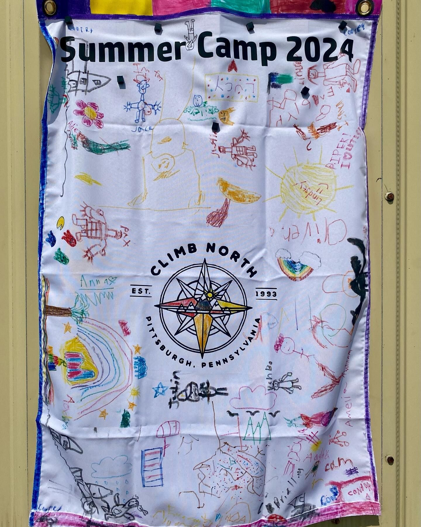 Summer Camp 2024 banner, white fabric, with children's drawings. Includes a compass rose logo for Climb North.