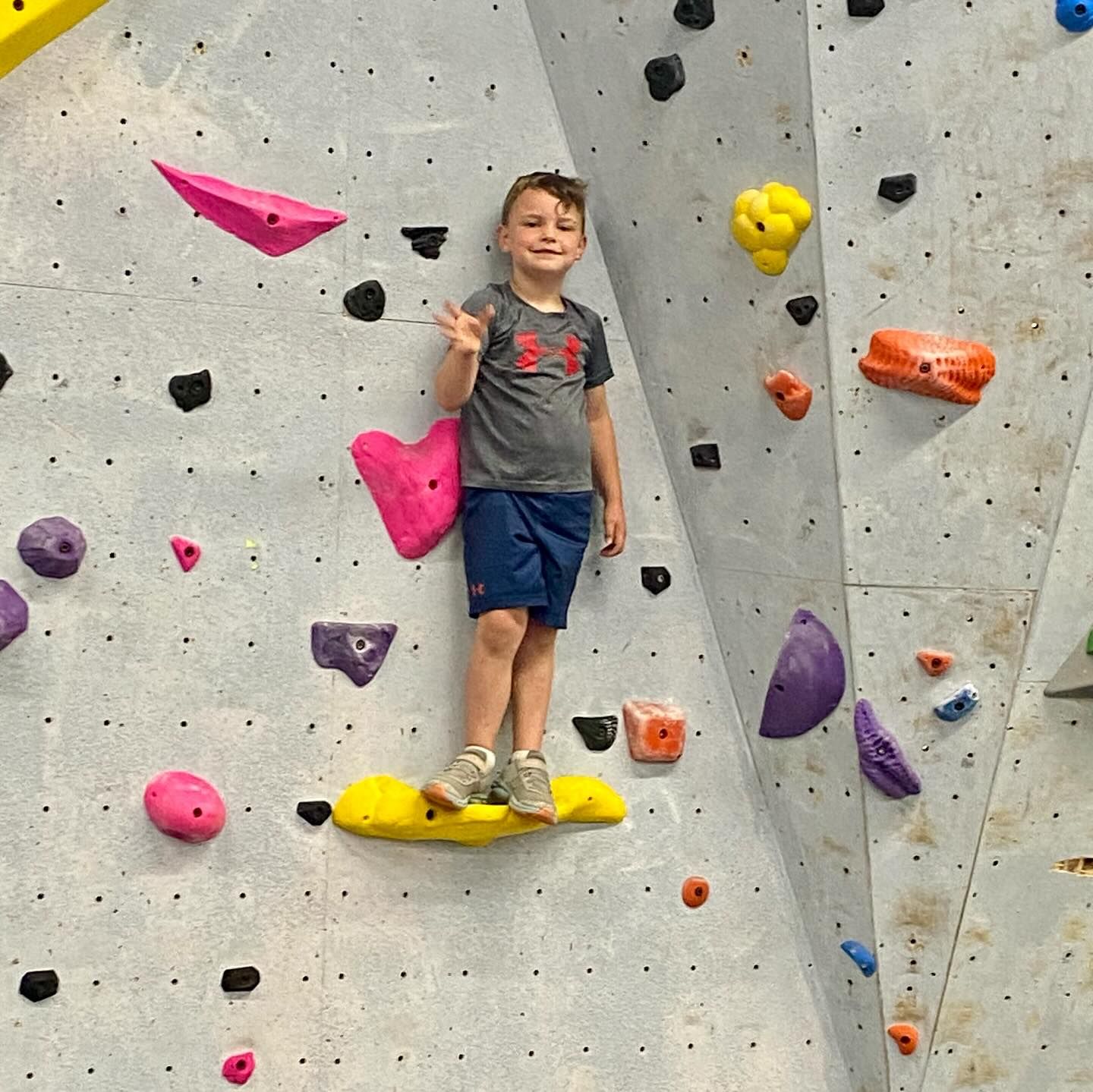 Boy on climbing wall, waving. Gray shirt, blue shorts, colorful holds.