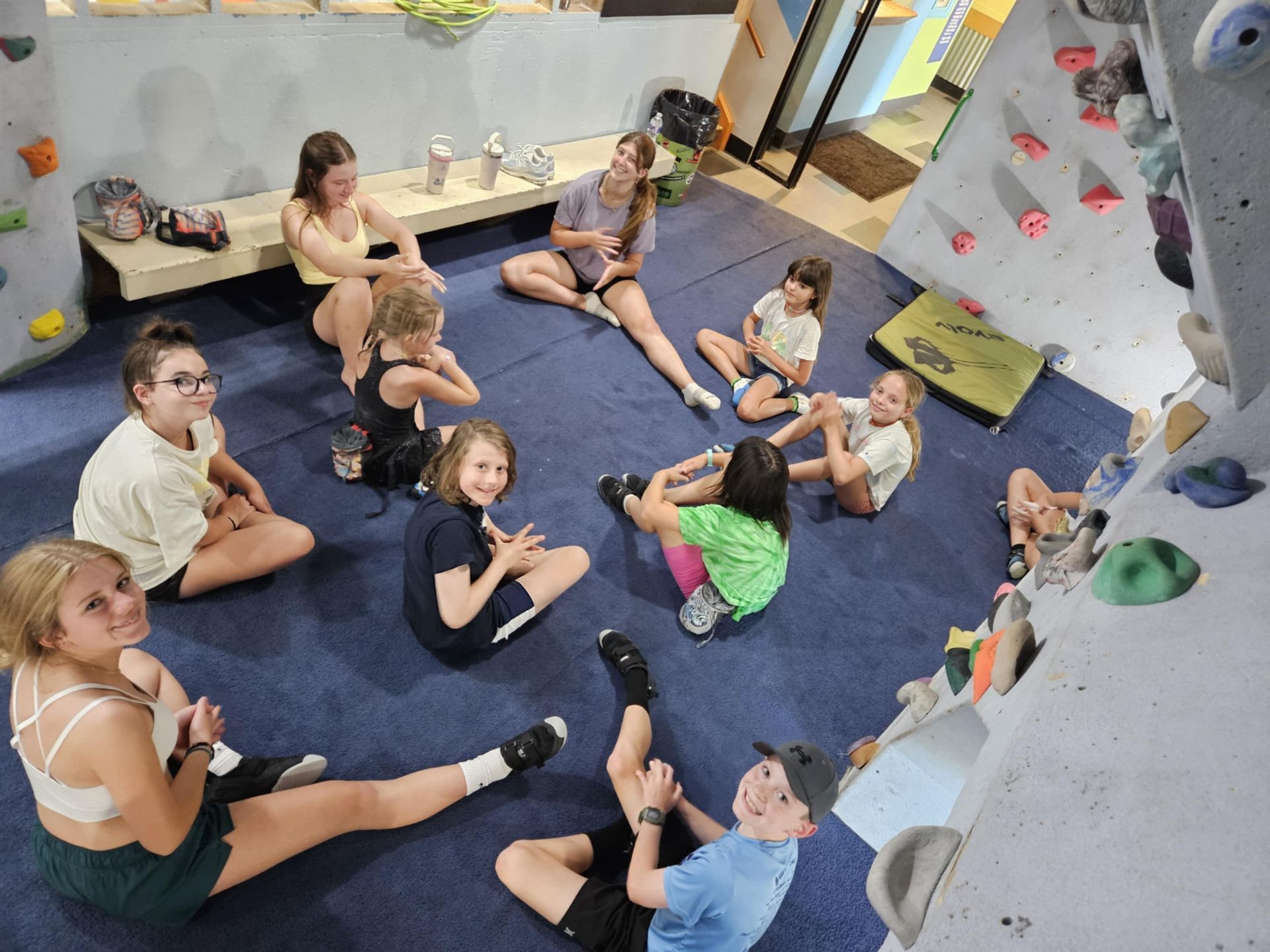 Group of kids sitting on a blue mat in a climbing gym, smiling.