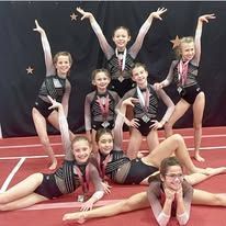 Young gymnasts in leotards posing together, some holding their arms up, others doing the splits.