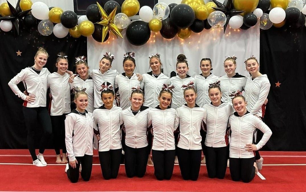Group of cheerleaders in black and white uniforms pose on a red mat, decorated with black and gold balloons.