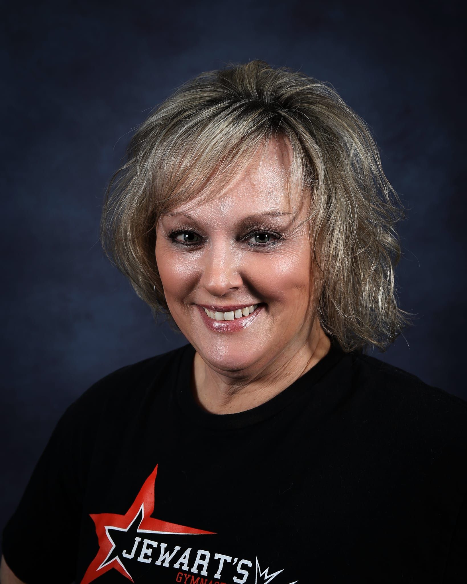 Woman with blonde hair smiling, wearing a black t-shirt with red star and white lettering, against a dark blue background.