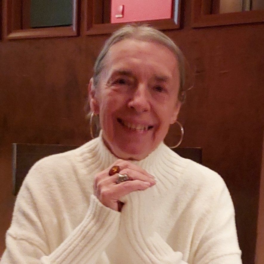 Smiling older woman in white sweater, hands clasped, wearing hoop earrings and rings.