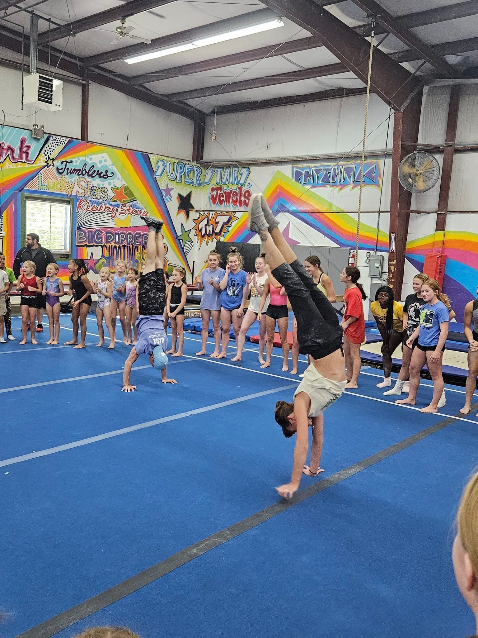 Two gymnasts doing handstands on a blue mat in a gym, while others watch.