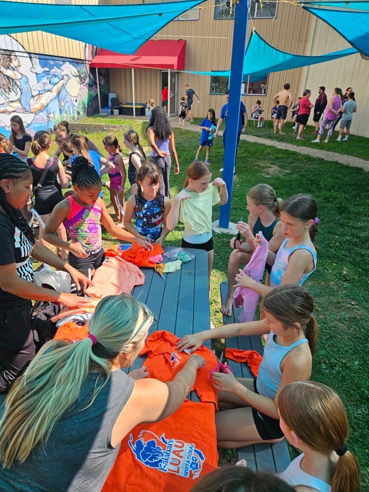Group of children with adults at picnic tables, with orange shirts, outside a building with a blue canopy.