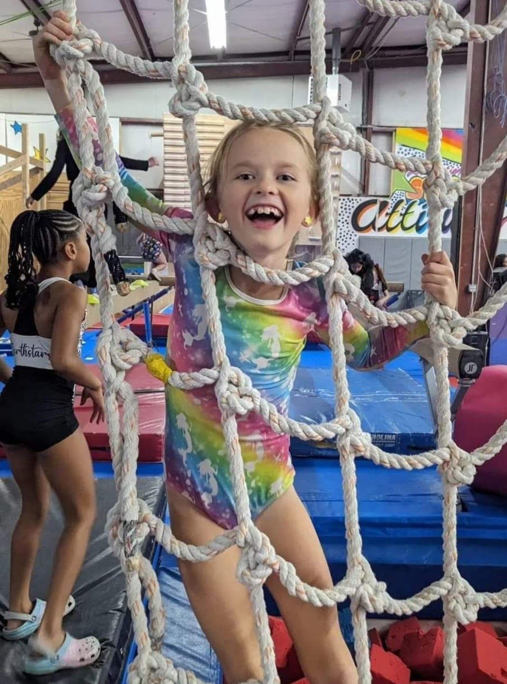 Girl in rainbow leotard smiling through climbing net at a gymnastics gym.