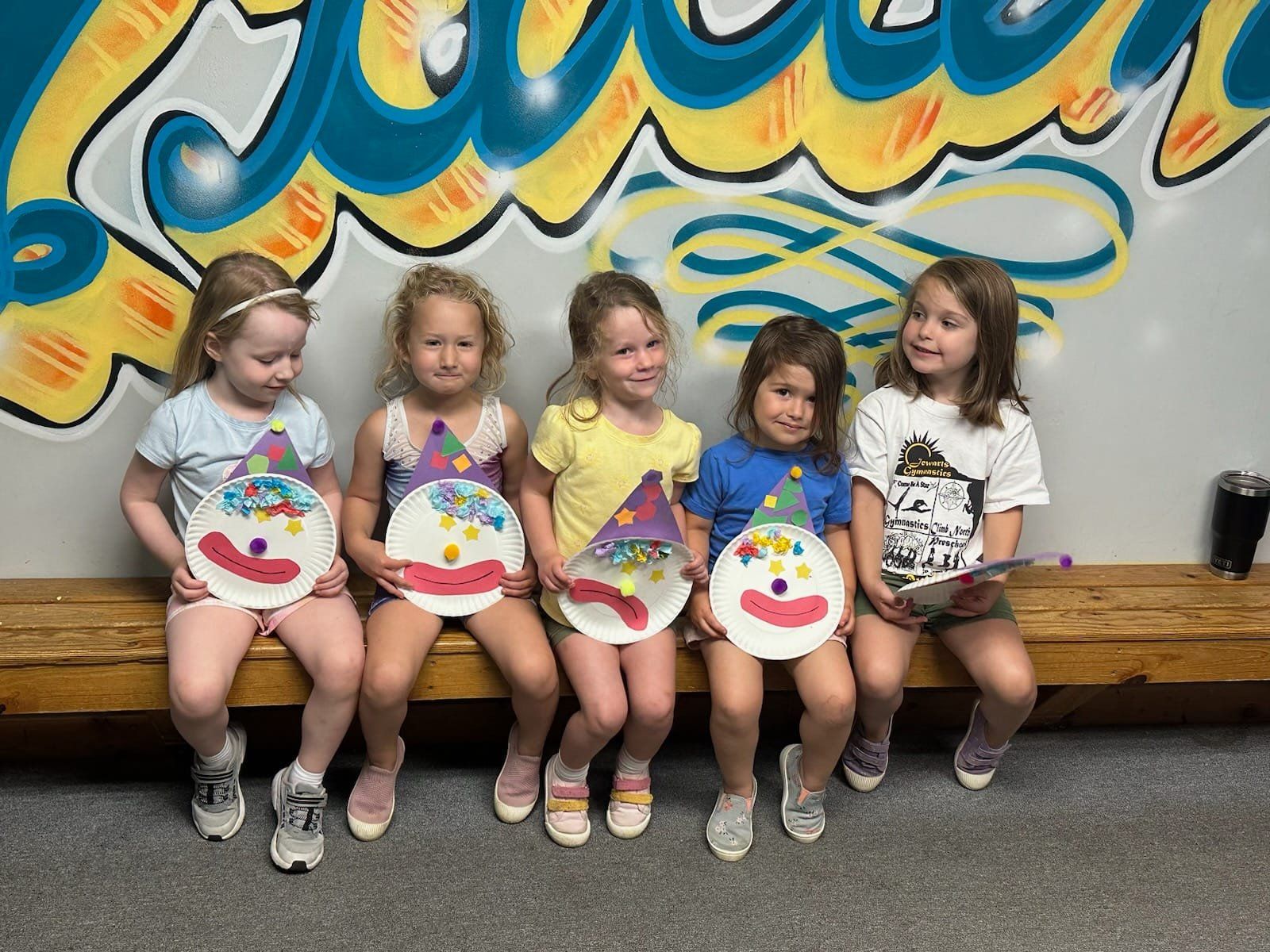 Five young girls holding paper plate clown crafts, smiling, sitting on a wooden bench in front of graffiti.