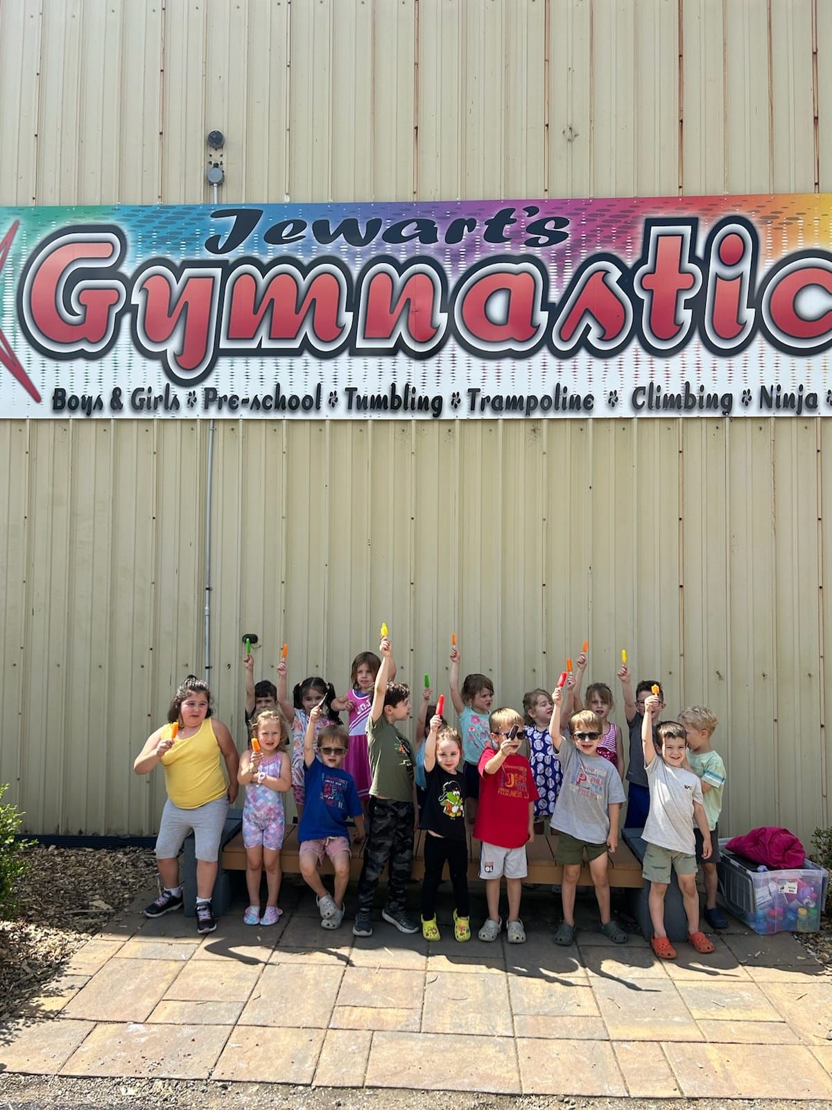 Children stand in front of a gymnastics sign, holding up colored sticks, outside.