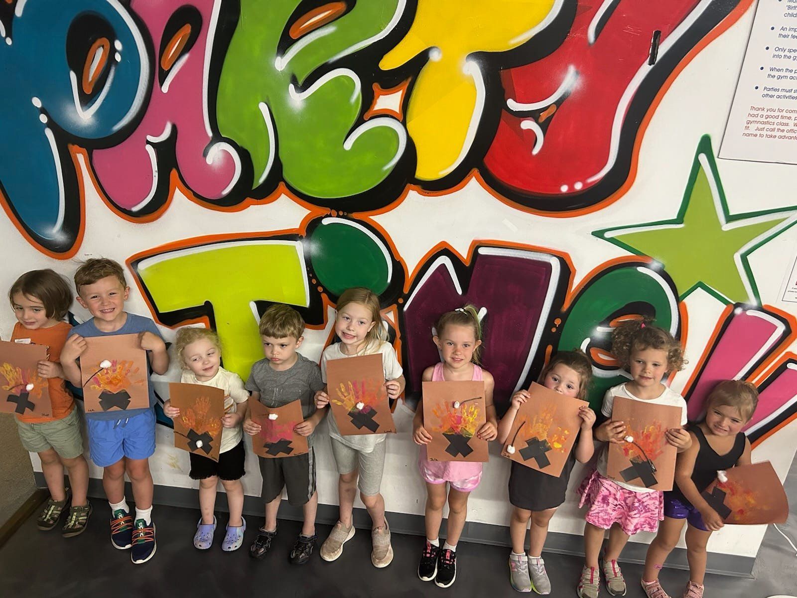 Children holding art projects in front of colorful graffiti.