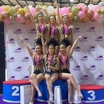 Group of gymnasts holding trophies, smiling, in front of a banner, after a competition.
