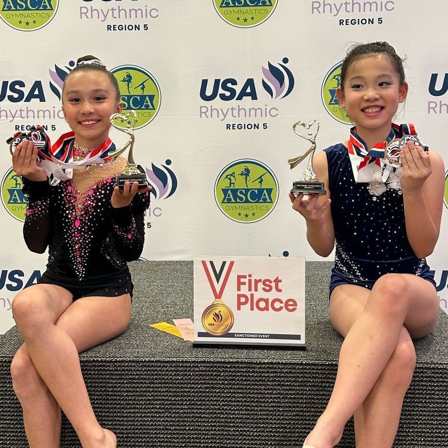 Two young girls in gymnastics outfits smiling, holding trophies and medals at a competition.