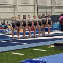 Gymnasts in leotards standing on a blue mat in an indoor gym.
