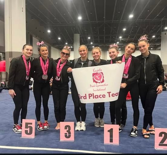 Gymnastics team celebrates 3rd place, holding a banner, wearing medals, and standing on numbered platforms.
