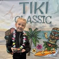 Young girl at a Tiki Classic event, holding a small tray. Smiling, in front of a tiki and palm tree.