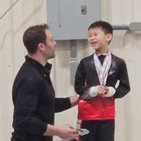 Man presenting a medal to smiling boy in a red and black uniform. Indoors, white background.