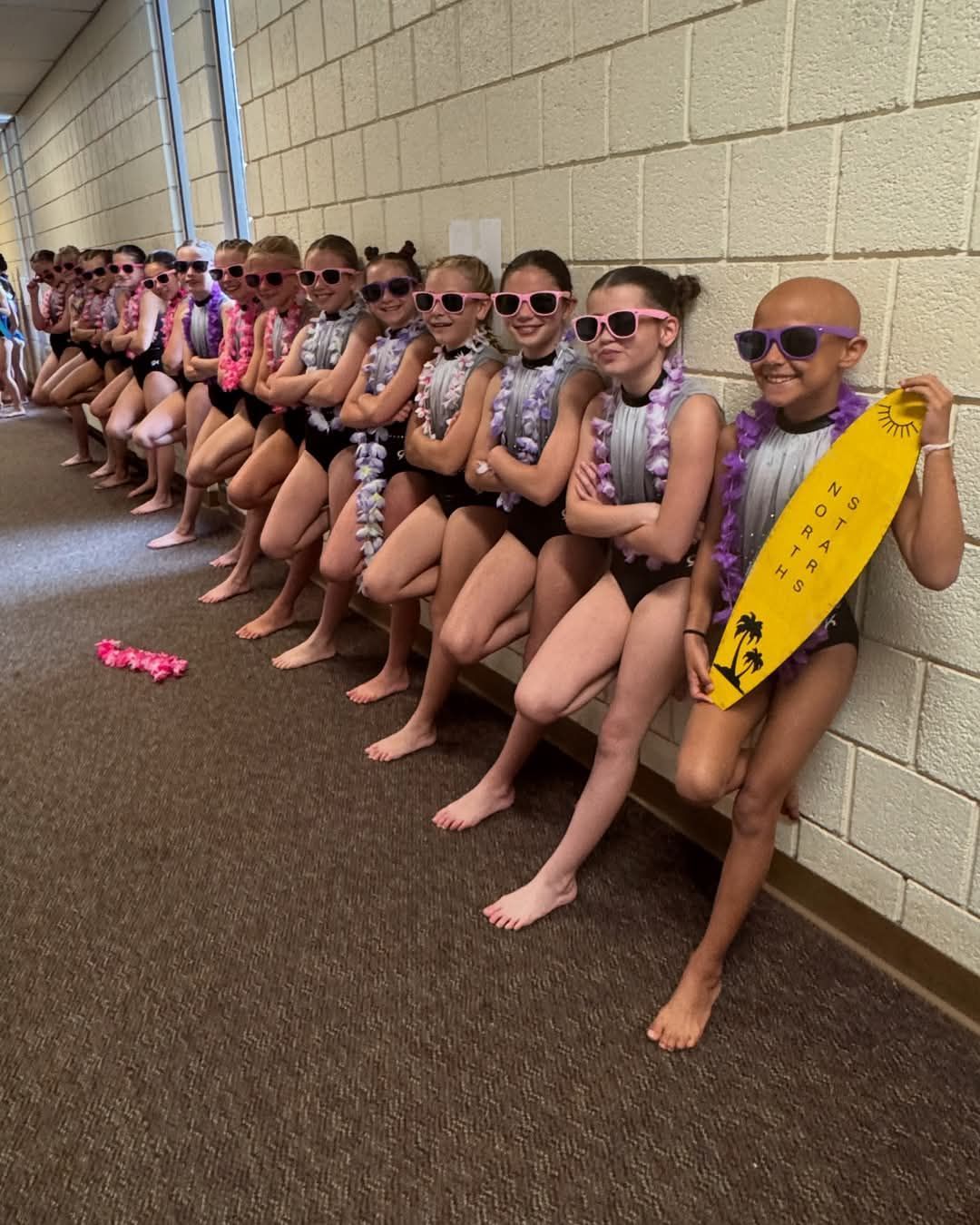 Row of young dancers in swimsuits and sunglasses, leaning against a wall. One holds a surfboard.