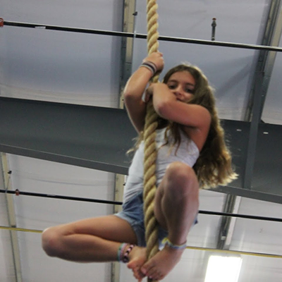 Girl climbing a rope indoors, wearing shorts and a tank top, smiling.